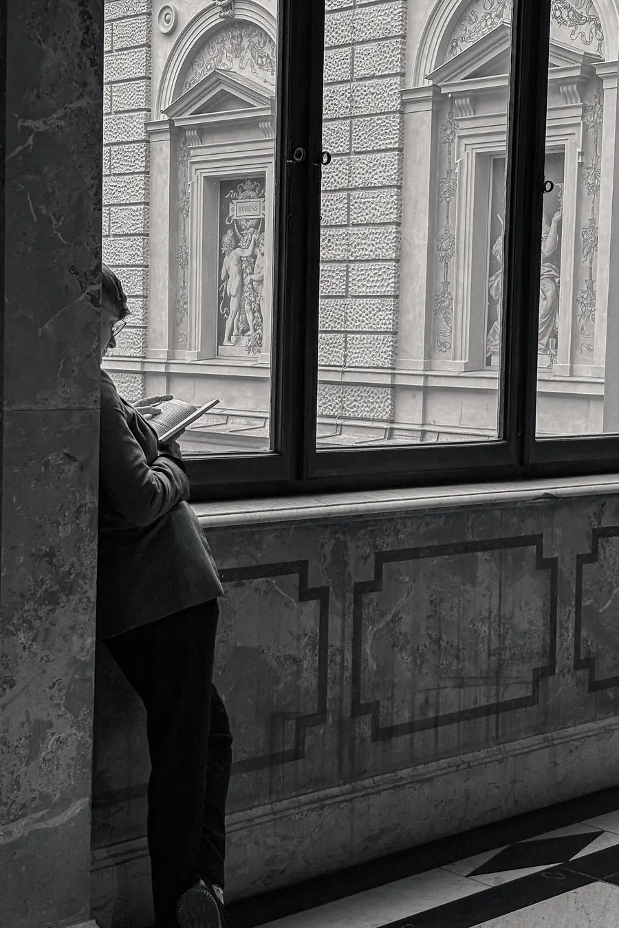 Figure reading by a museum window, ornate facade opposite