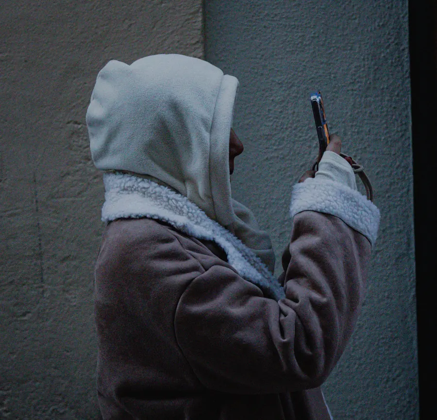 Hooded figure photographing with a phone against a pale wall