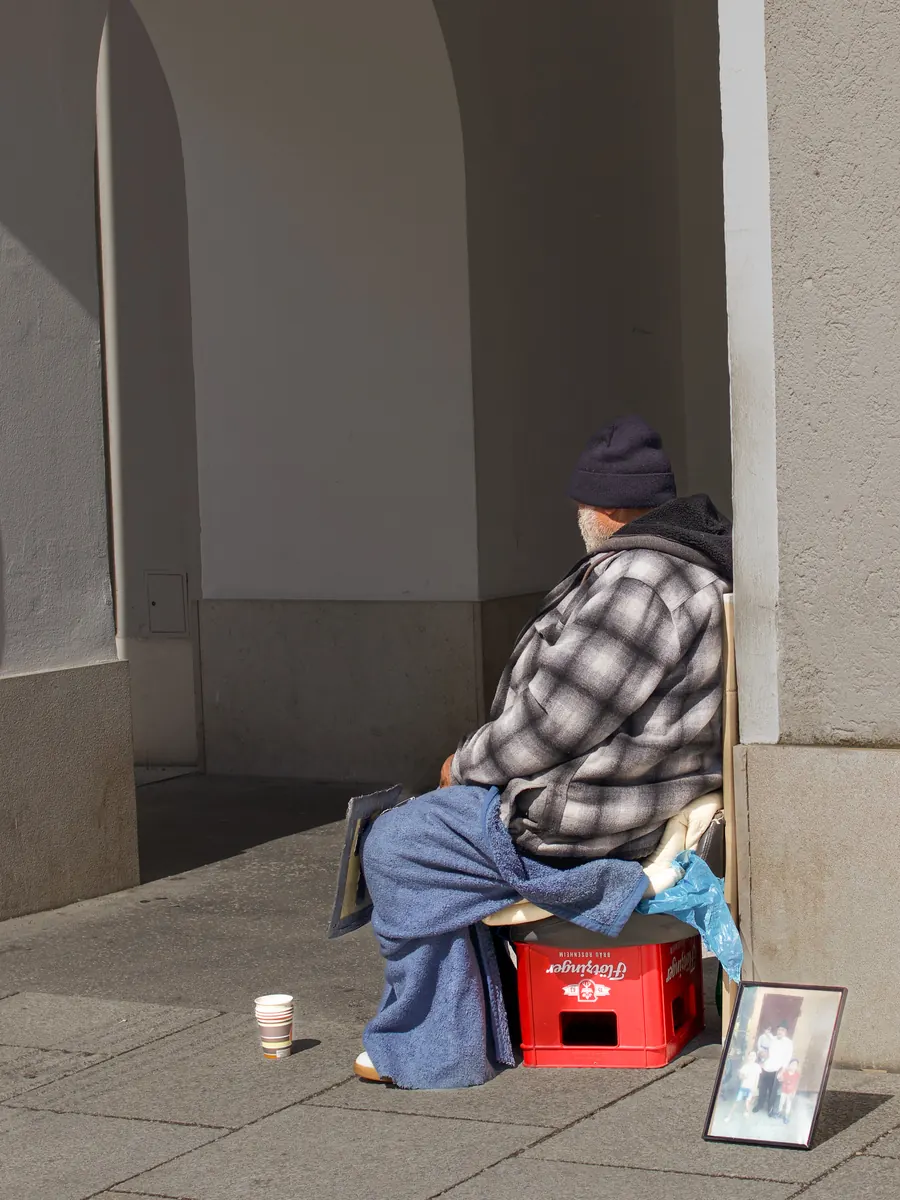 Man seated on a red crate under an archway, a cup and a framed photo beside him