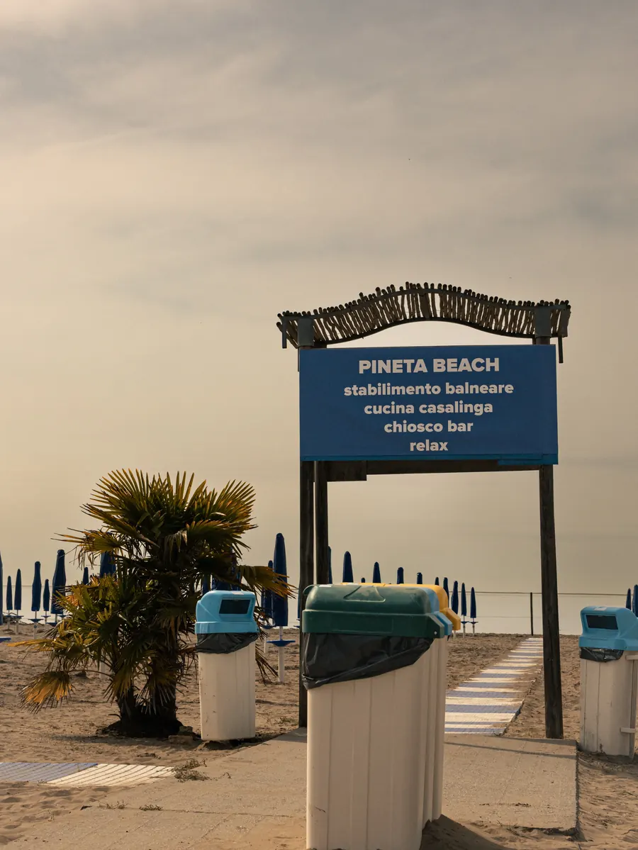 Pineta Beach sign above bins and a palm, umbrellas folded behind